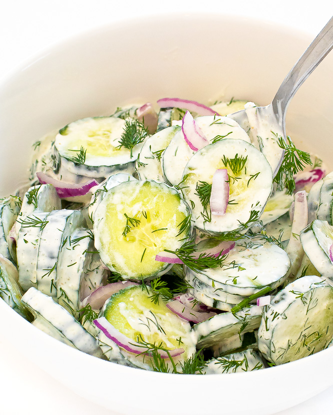 Creamy cucumber dill salad in a white bowl with a silver spoon scooping some out. 