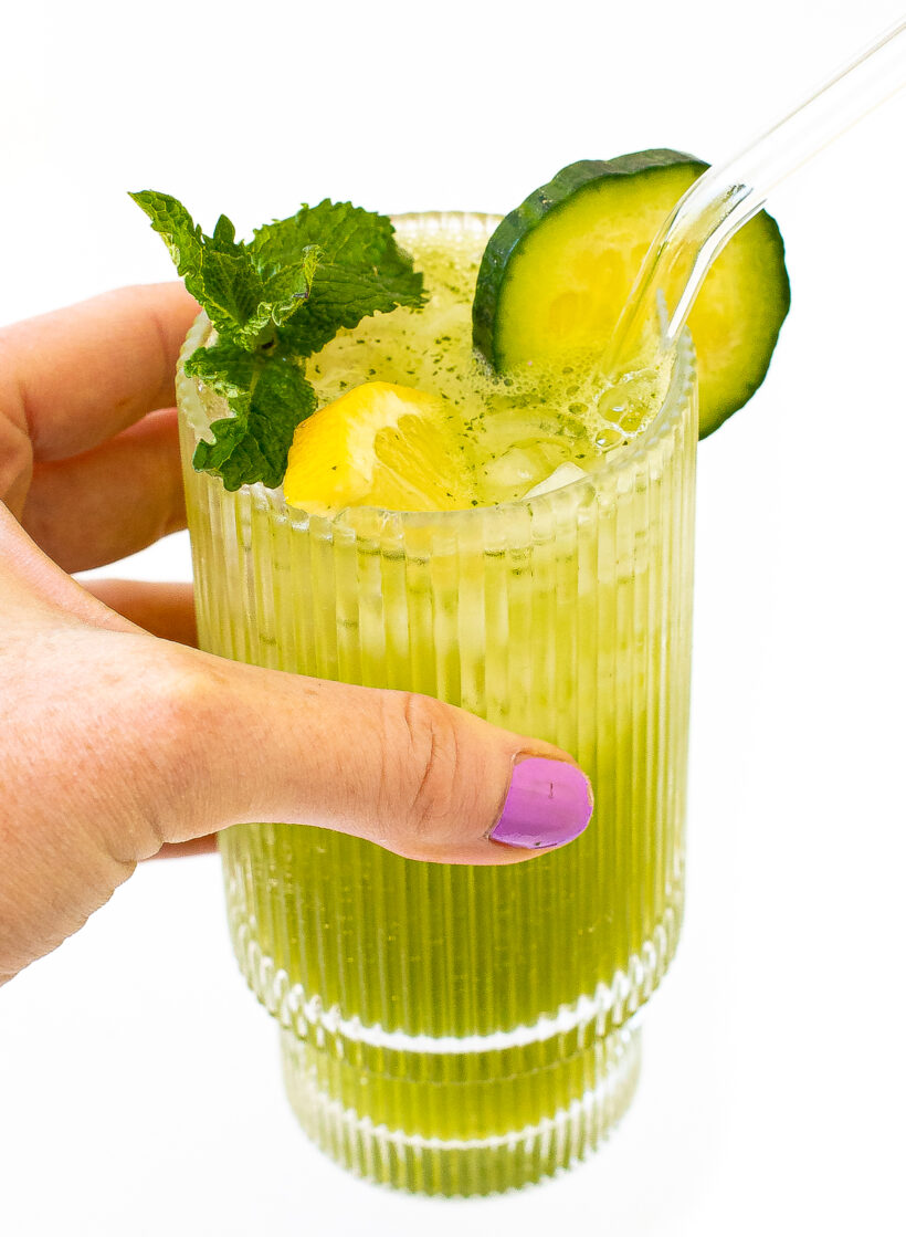 Hand holding a glass of lemonade garnished with mint, cucumber and lemon with a glass straw.