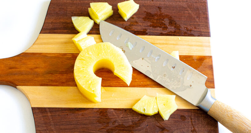 Pineapple rings being chopped on a wooden cutting board with a large knife.