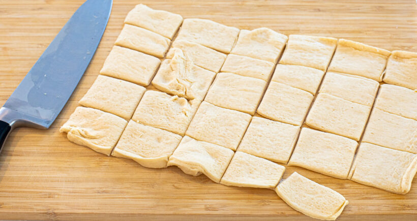 Crescent roll dough cut into squares on a wooden cutting board. 