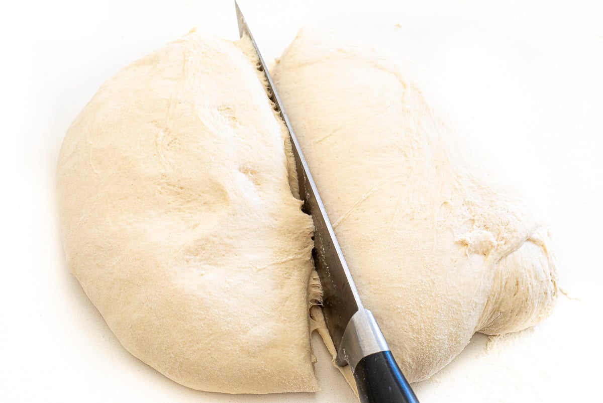 A knife slicing through a ball of sourdough bread dough on a white surface.