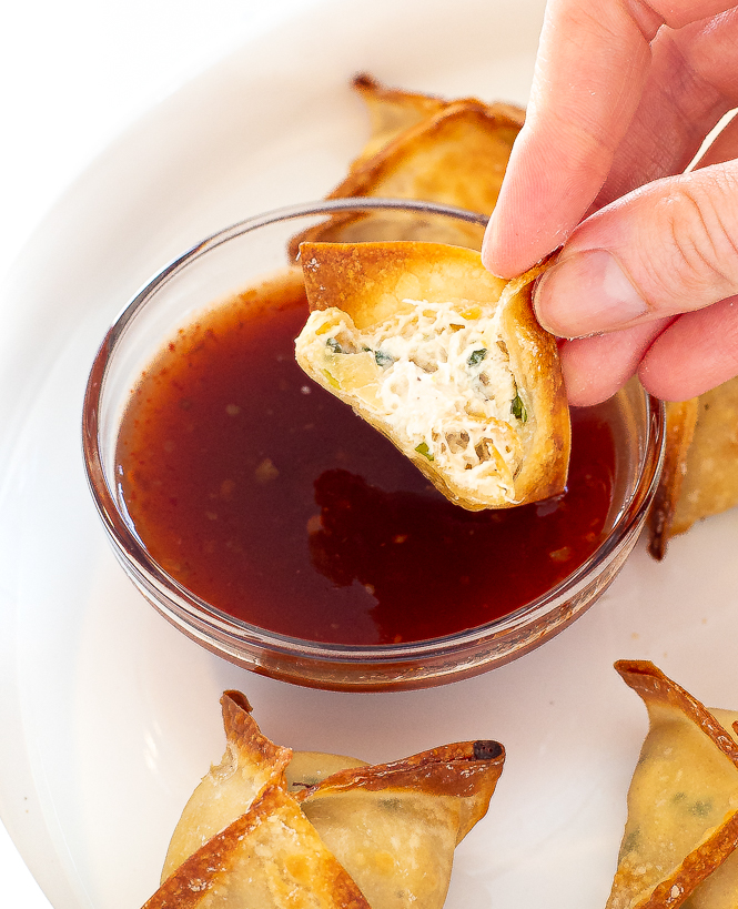 Hand holding a crab rangoon that is missing a bite with sauce in the background. 
