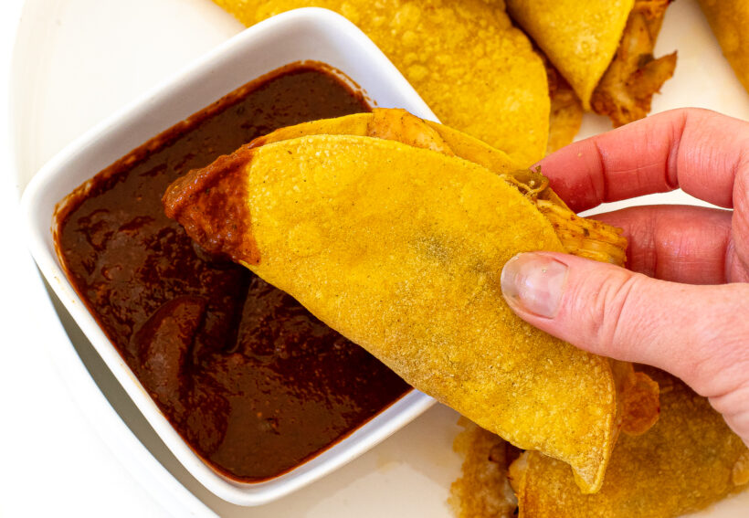 A hand holding a crispy baked chicken taco and dipping it in the chili salsa.