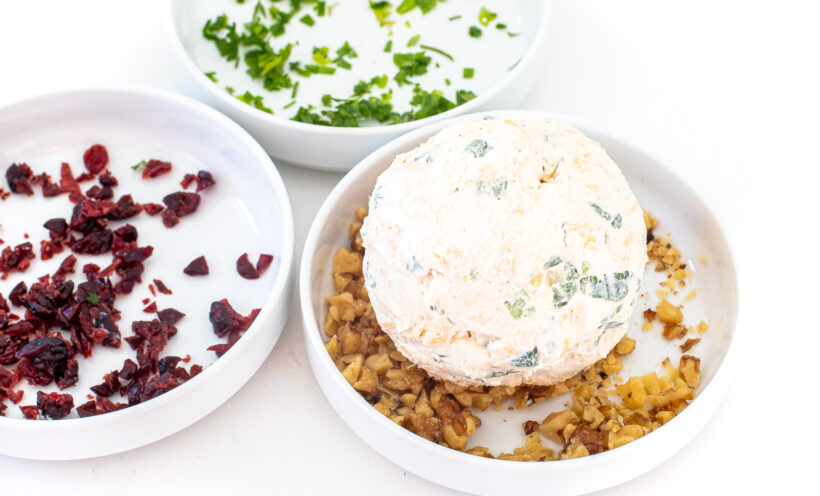 Cheeseball in a bowl with walnuts. Bowls with cranberries and parsley are in the background.