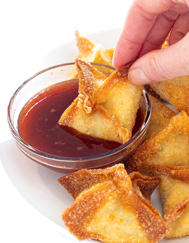 Hand holding a crab rangoon and dipping it into a red sauce. 