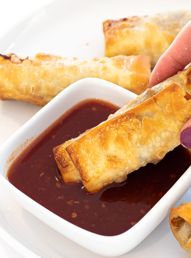 Egg roll being dipped into a red dipping sauce by a hand. 