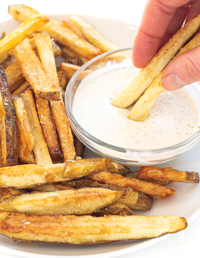 Hand dipping two french fries into small bowl ranch sauce.