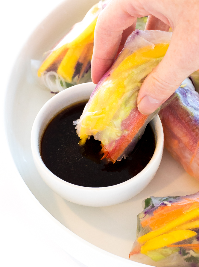 the author holding a Rainbow Vegetable Spring Roll and dipping it into the sauce with more rolls on a white plate