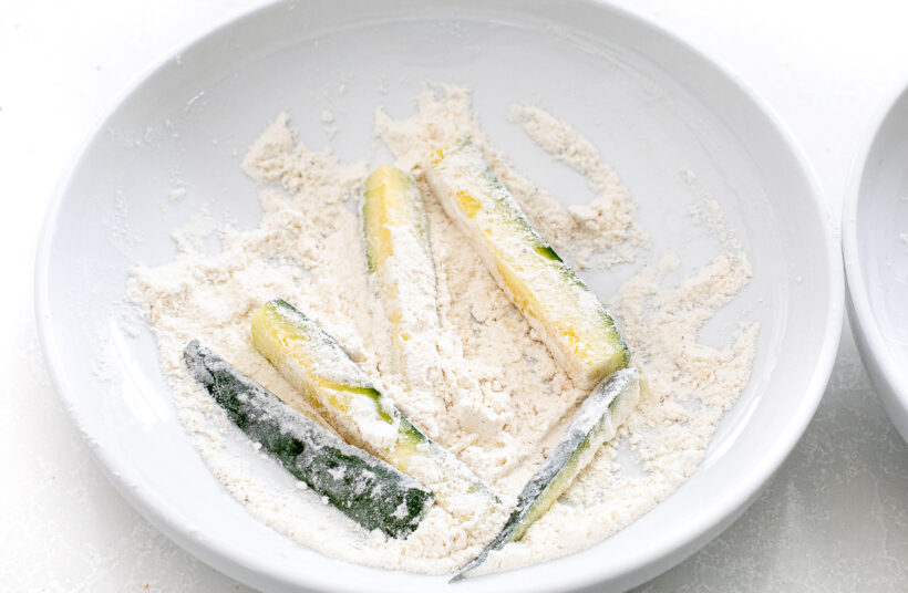 Zucchini being dredged in flour in a small bowl.