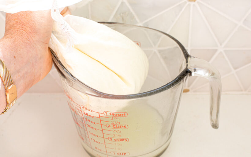 A hand holding a nut bag with curd mixture draining into a glass measuring cup.