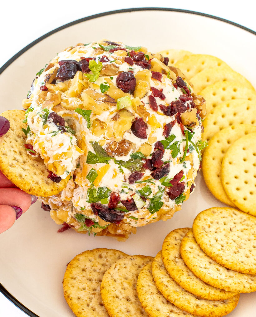 A cheese ball on a plate with crackers. A hand is dipping a cracker into the cheeseball.