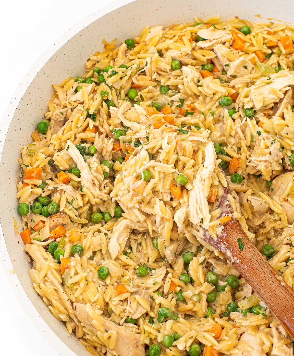 A wooden spoon scoops a serving of creamy chicken, orzo pasta, and melting potatoes with peas and carrots from a large pan.