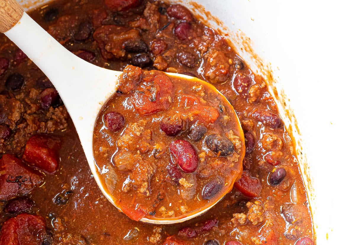 A white ladle scoops up hearty Slow Cooker Chili with ground beef, kidney beans, black beans, and tomatoes from a pot.