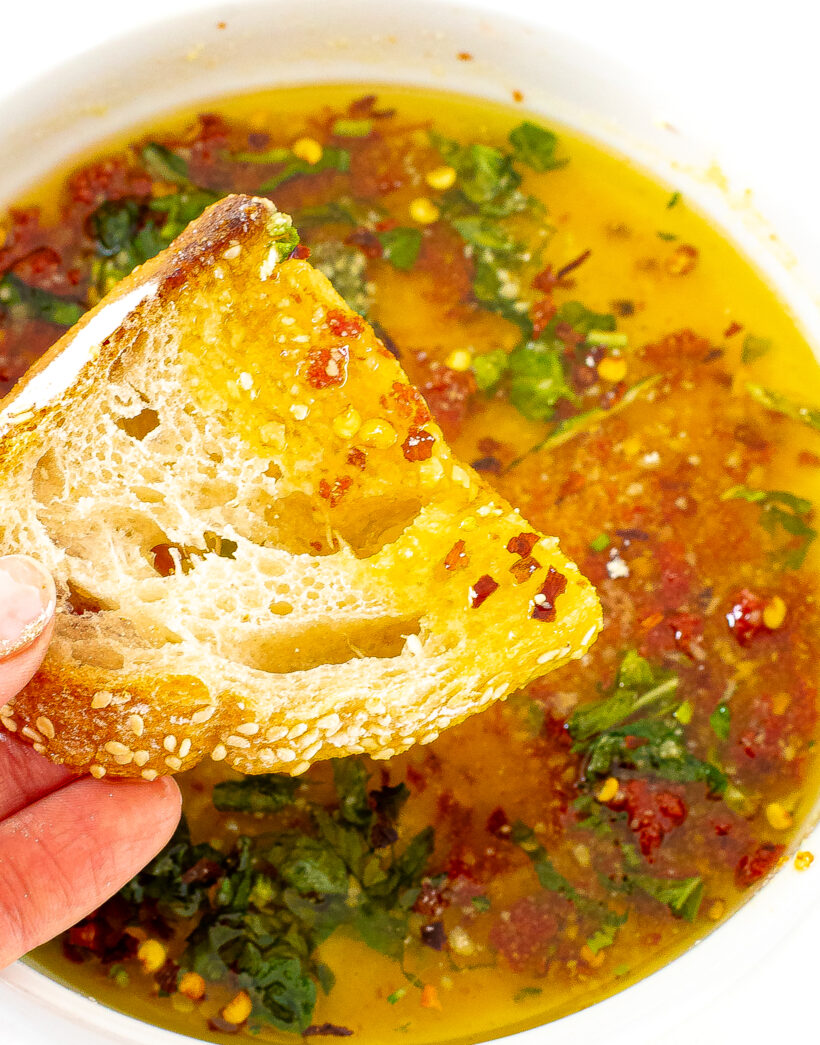A hand holding a piece of bread with dipping oil on it above the bowl with the rest of the oil.