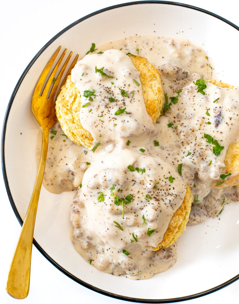 3 fluffy biscuits topped with sausage gravy on a white plate with a gold spoon.