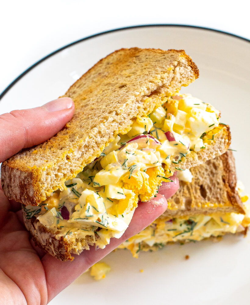 Hand holding half of an egg salad sandwich with the other half in the background. 