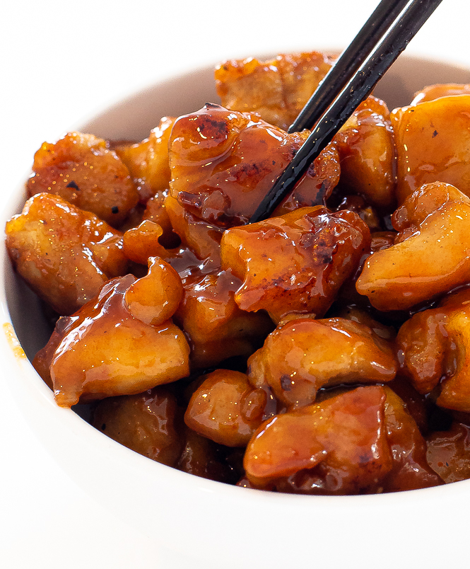 Chicken coated in a sauce in a white bowl with chopsticks grasping a piece of chicken.