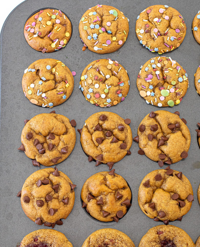Mini flourless peanut butter muffins in a muffin tin with some of them topped with chocolate chips and others topped with sprinkles. 
