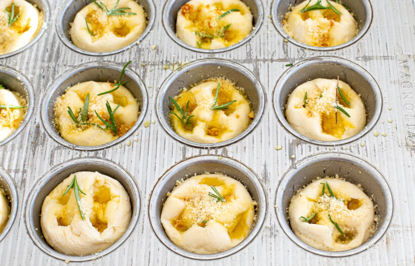 Focaccia dough in a muffin tin topped with olive oil, parmesan and rosemary before being baked. 