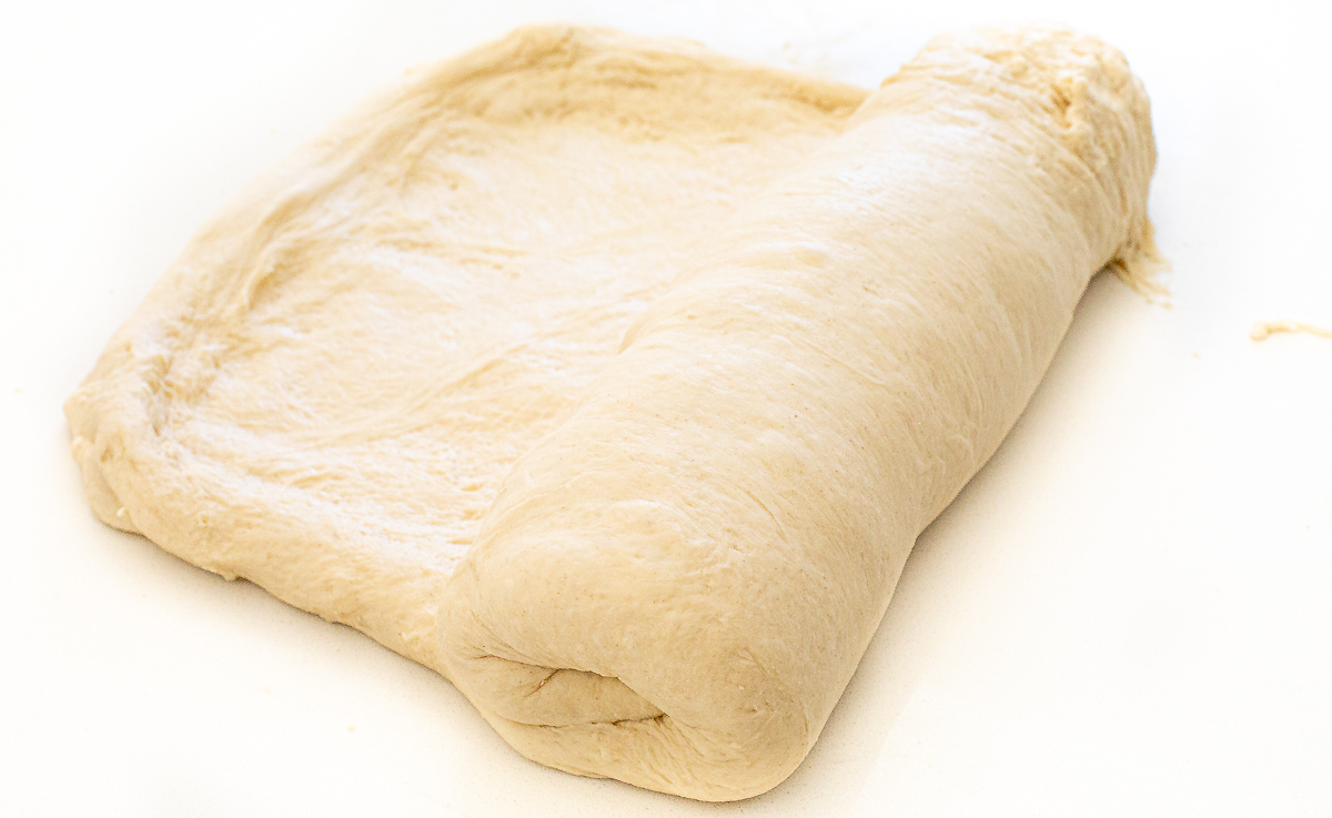 A rectangular sheet of raw sourdough sandwich bread dough being rolled into a cylinder.
