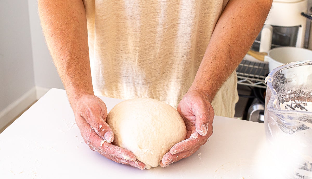 Hands shaping a round ball of sourdough sandwich bread dough on a white surface.