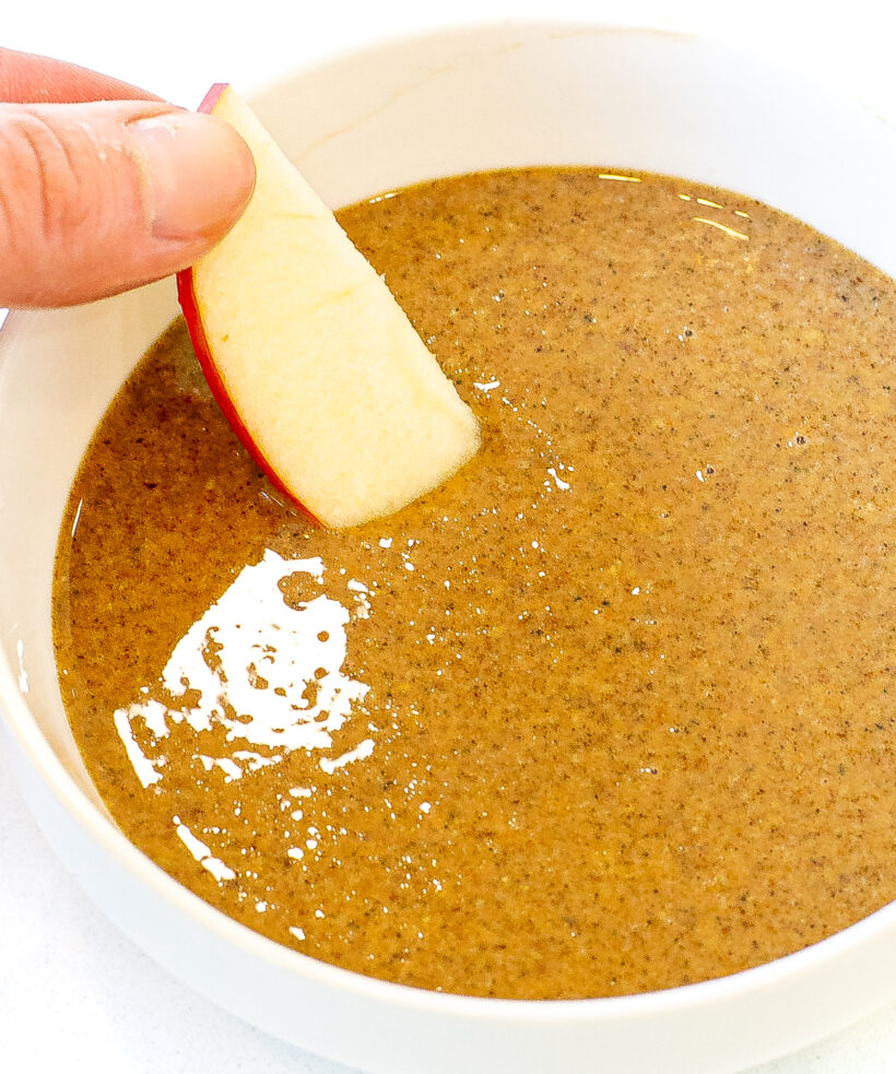 Hand dipping an apple slice into the granola butter in a bowl. 