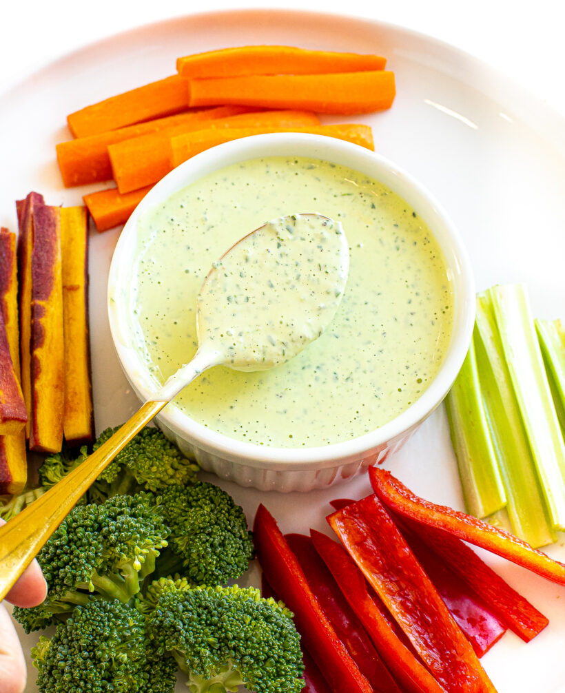 Spoon holding up green goddess dressing over a white bowl with veggies around it.