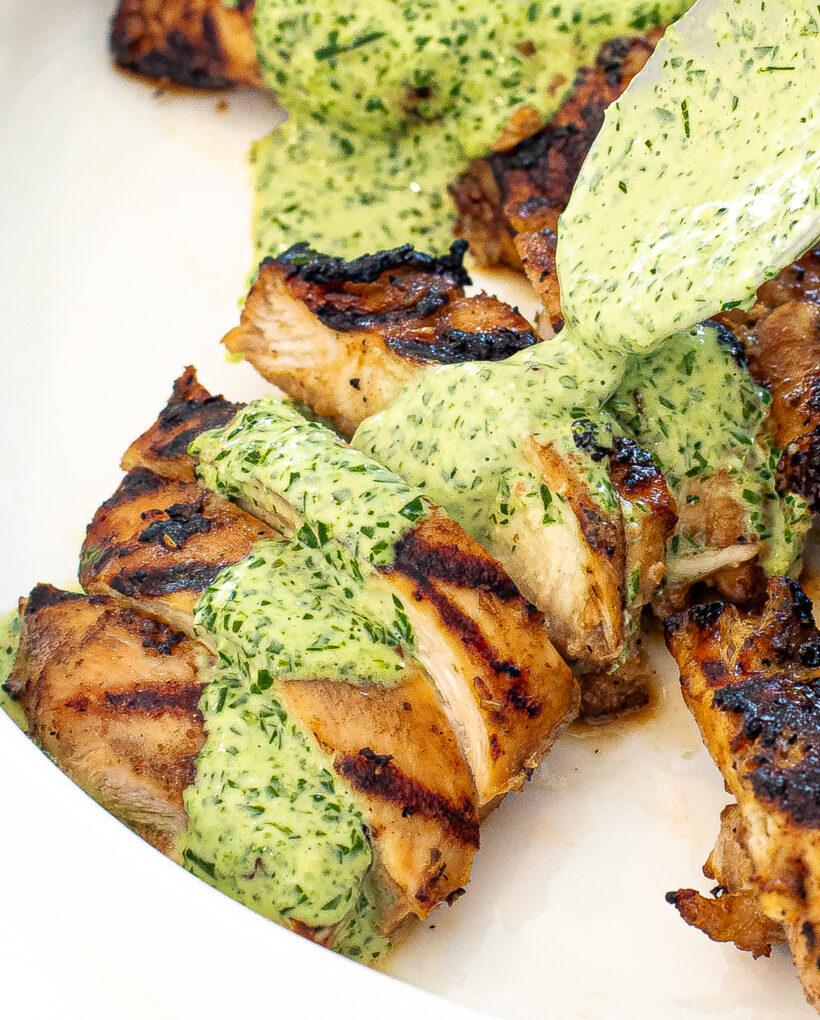 Grilled chicken sliced on a white plate with a green sauce being drizzled over top. 