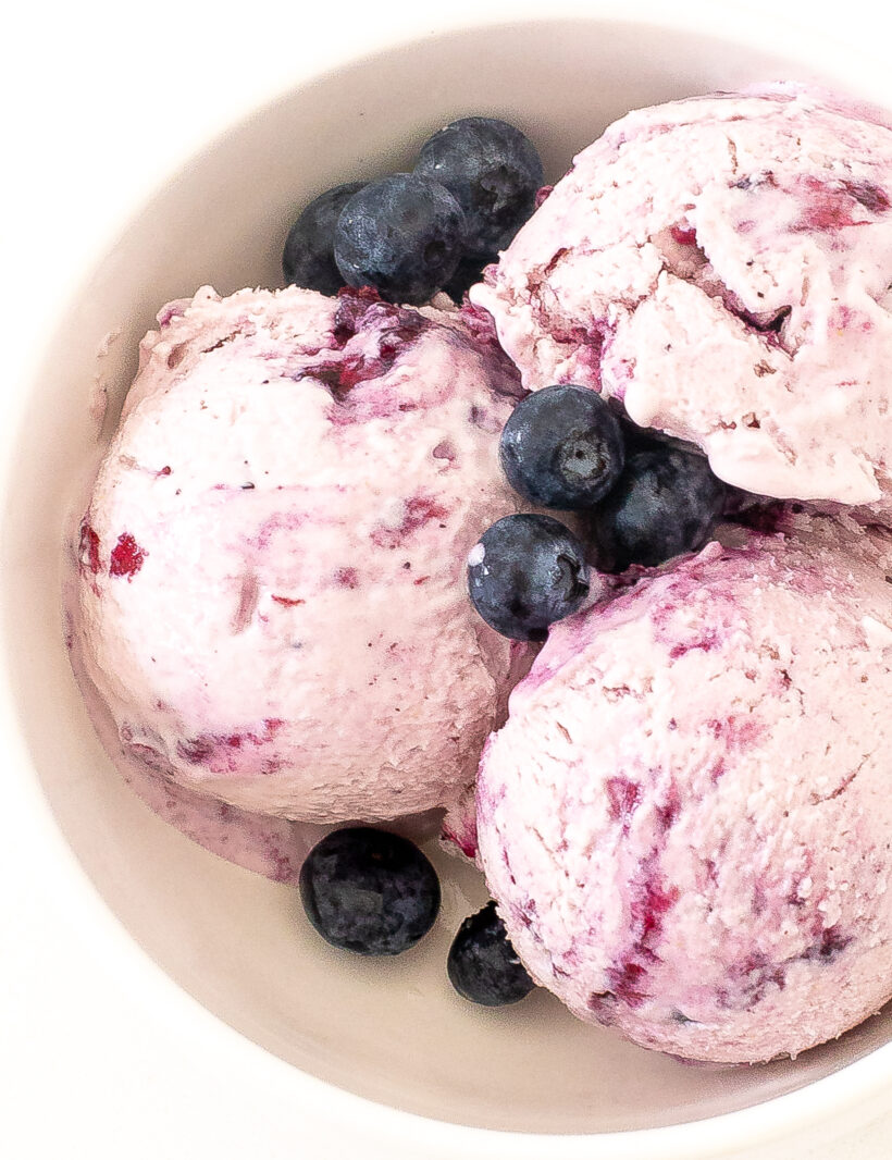 Three scoops of blueberry ice cream in a bowl with fresh blueberries on top. 