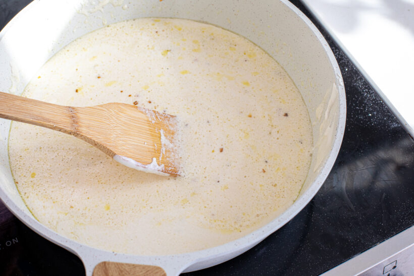A wooden spoon stirring alfredo sauce in a white skillet.