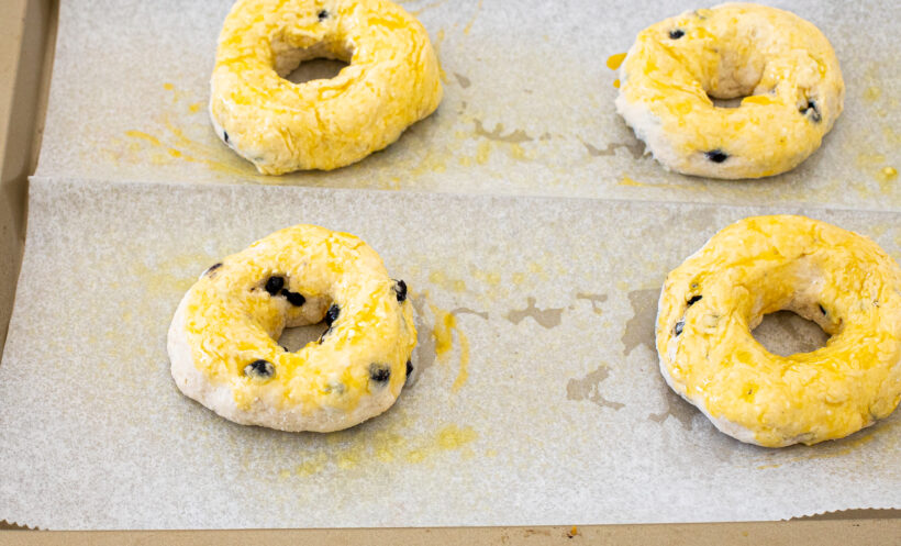 Blueberry bagels brushed with egg wash and placed on a baking sheet.