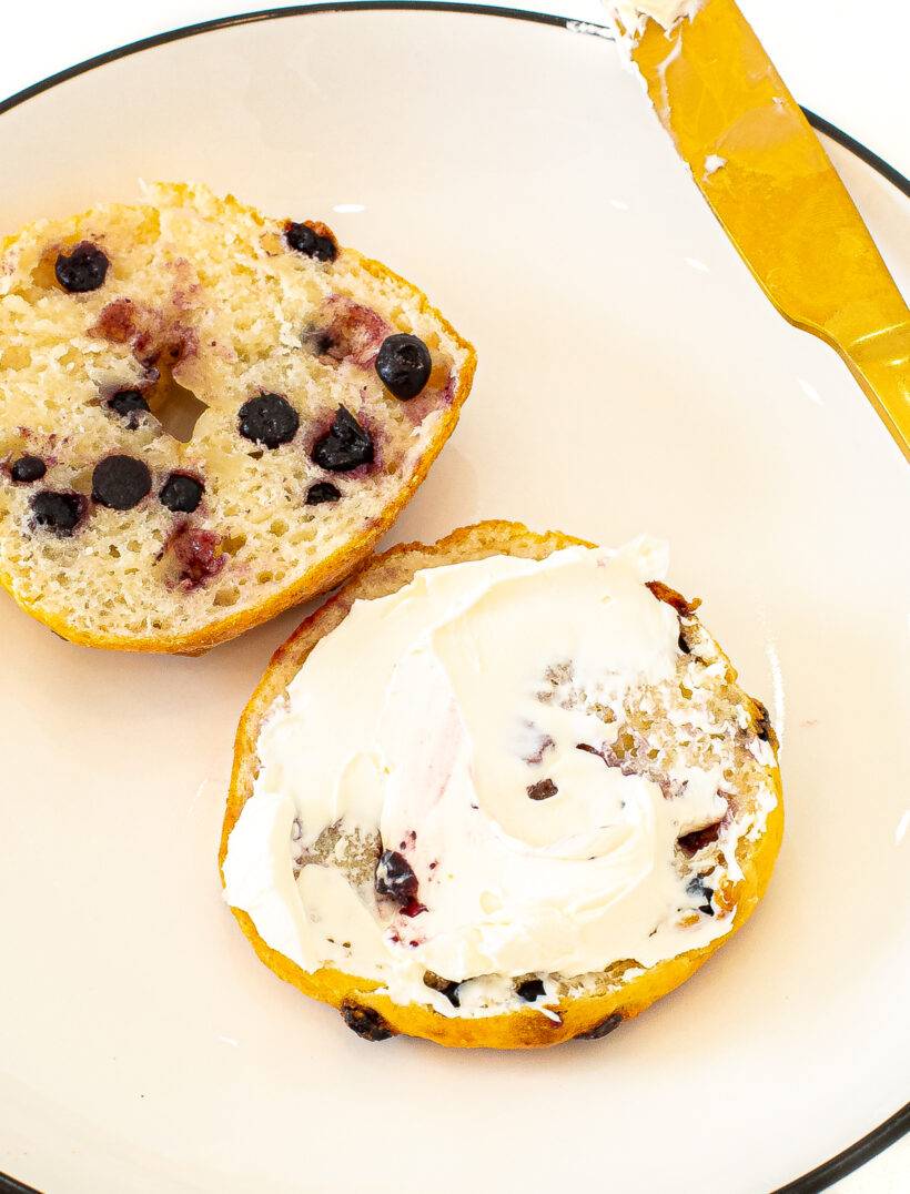 A blueberry bagel cut in half with one piece of the bagel smeared with cream cheese.