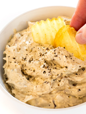 a white bowl filled with this caramelized onion dip and the author taking a scoop of it out with a potato chip