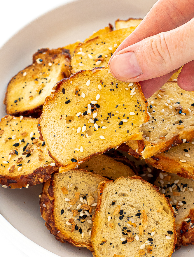 A hand holding a bagel chip with bagel chips in a white bowl.