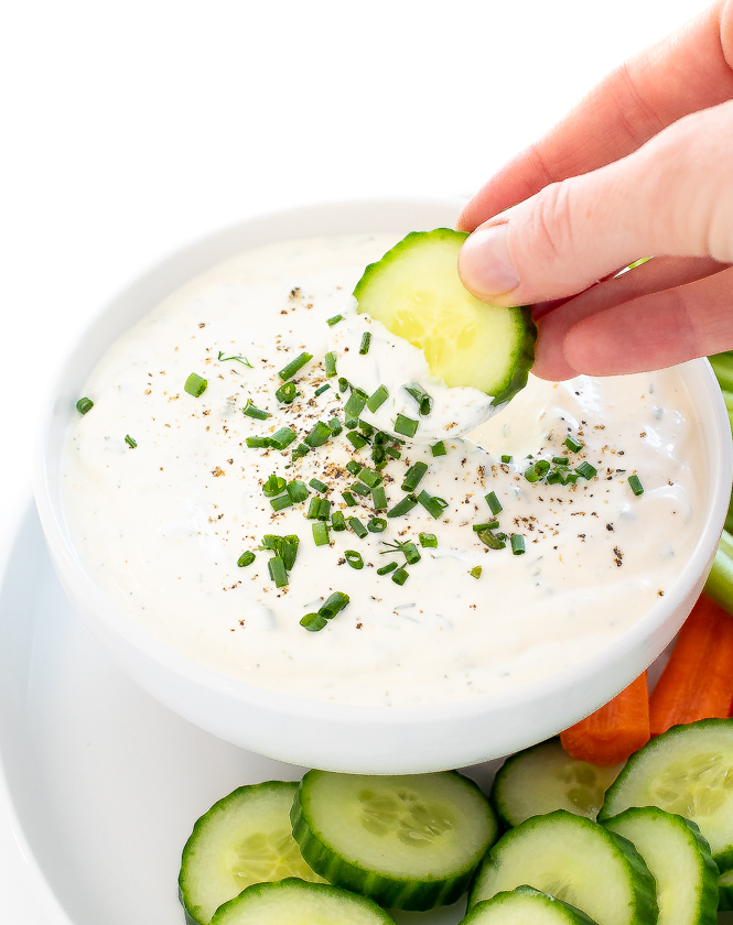 A cucumber being dipped into homemade ranch in a white bowl next to cucumber and carrots.