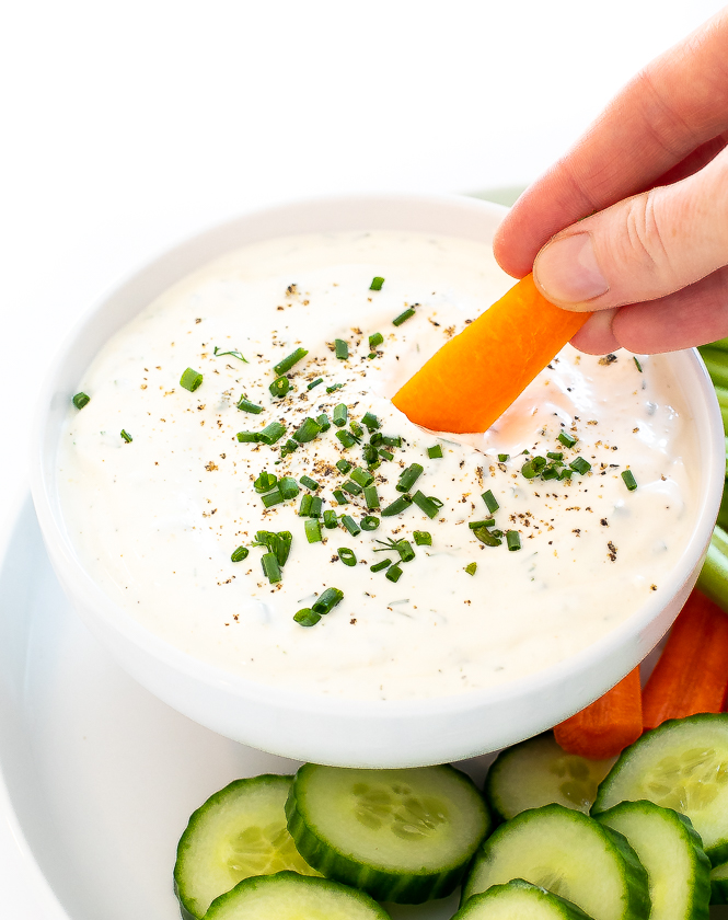 A carrot being dipped into homemade ranch in a white bowl next to cucumber and carrots.