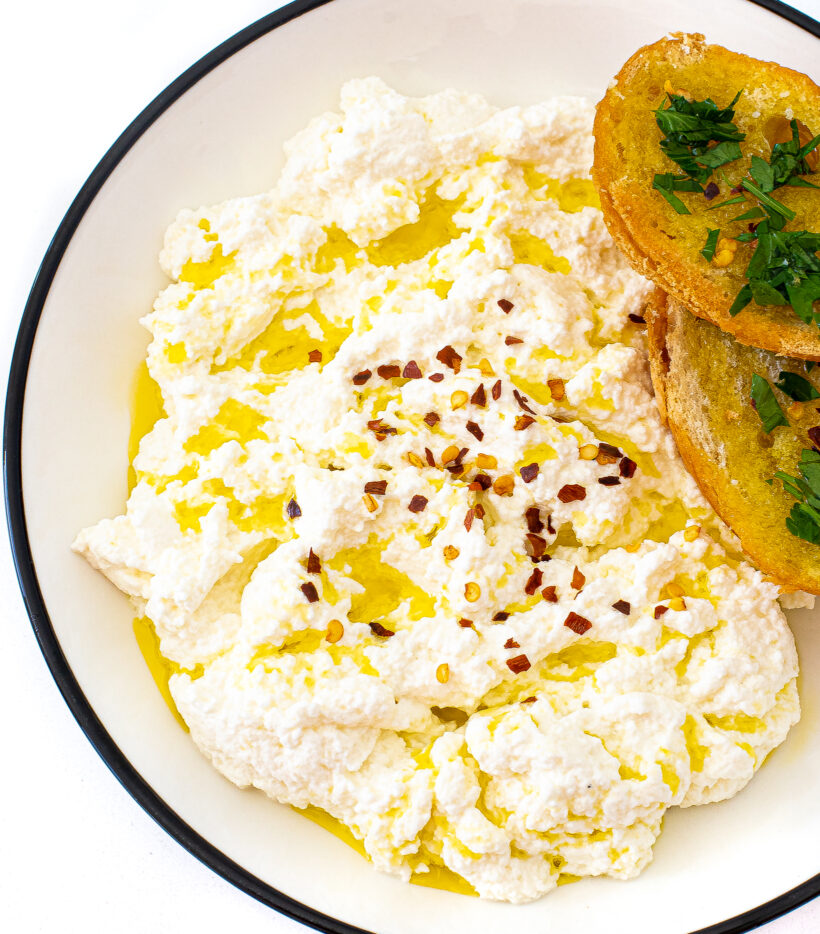 A bowl of ricotta topped with olive oil and red pepper flakes. Two pieces of crostini are in the bowl.