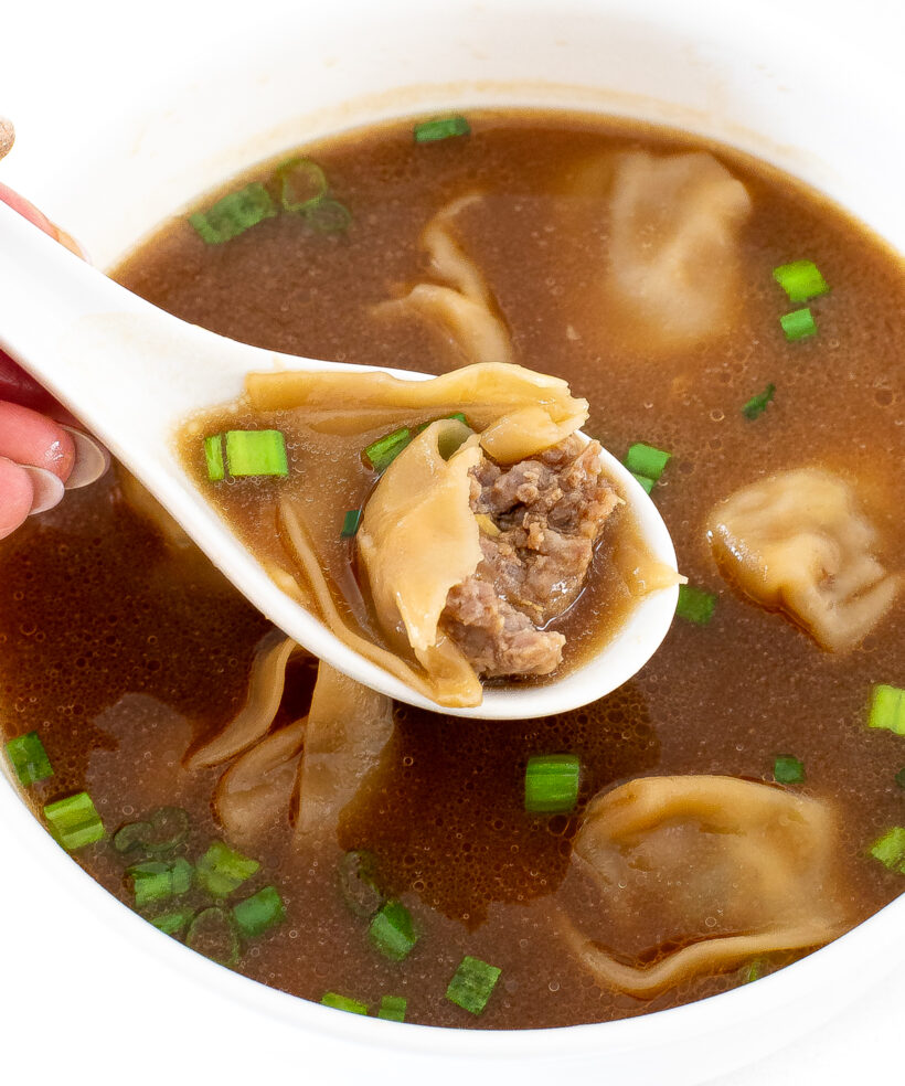 A hand holding a white spoon with a spoonful of wonton soup above a bowl of soup.
