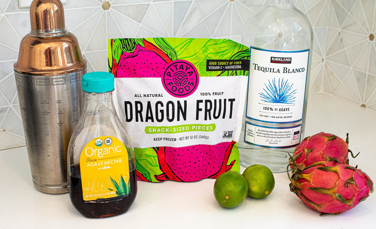 A bottle of tequila near frozen dragon fruit, limes, agave and a cocktail shaker.
