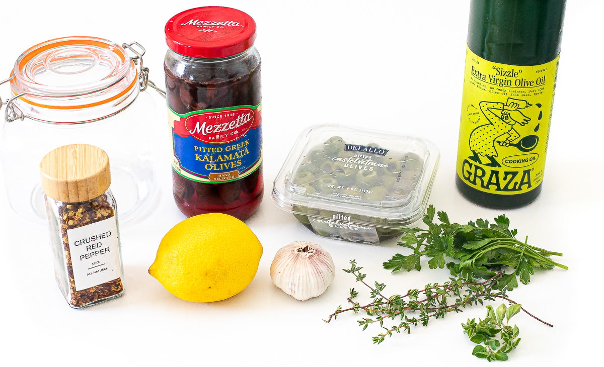 Fresh herbs near a lemon, jar of olives, garlic and olive oil.