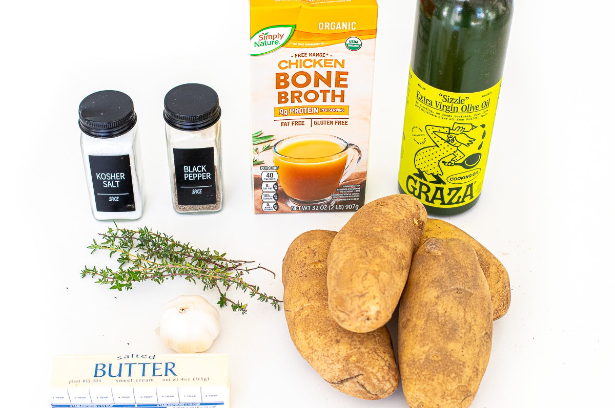 Ingredients for melting potatoes on a white surface: a carton of chicken bone broth, olive oil, salt, pepper, a head of garlic, fresh thyme, butter, and three russet potatoes.