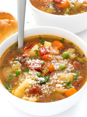 A bowl of easy vegetable soup topped with grated cheese and herbs, with a spoon inside. Another bowl and a bread roll are partially visible in the background, showcasing how to make vegetable soup at home for a comforting meal.