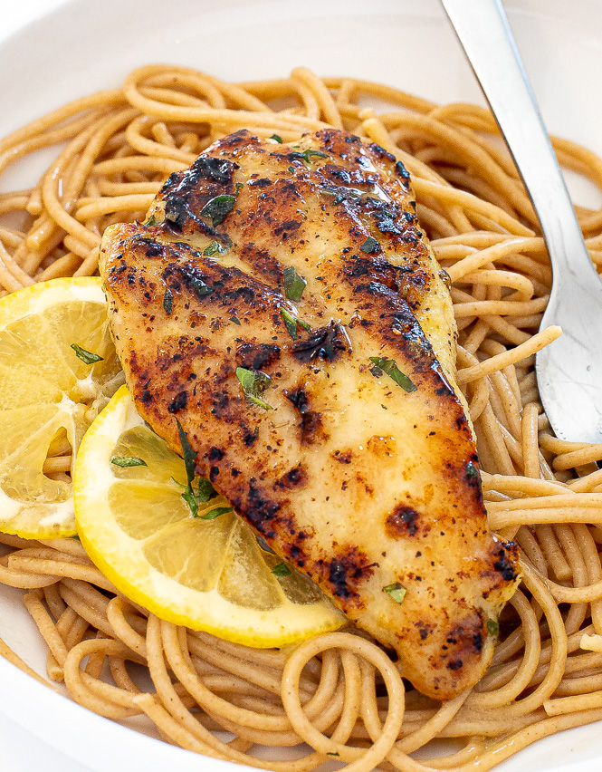 Piece of chicken on top of bed of pasta with lemon slices on top. 