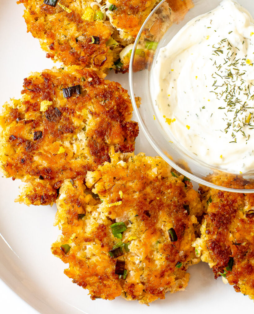 Close up view of salmon cakes on a white serving plate next to a bowl of lemon dill sauce.
