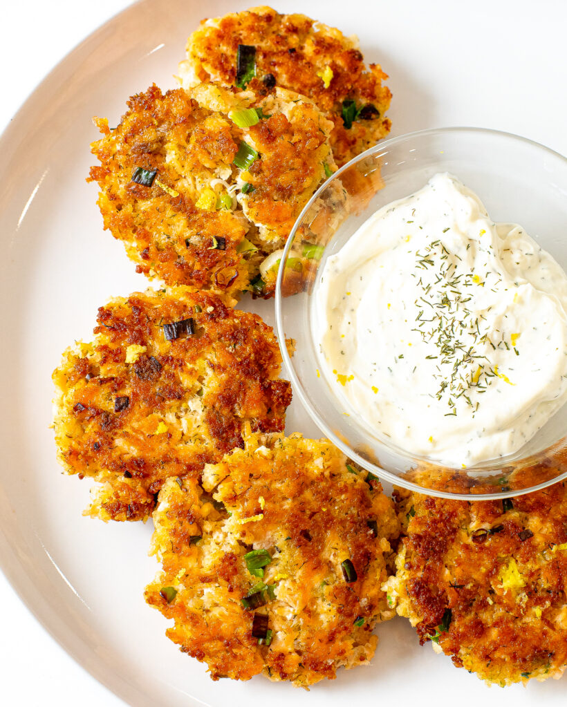 Salmon cakes laid out on a white plate next to a bowl of lemon dill sauce.