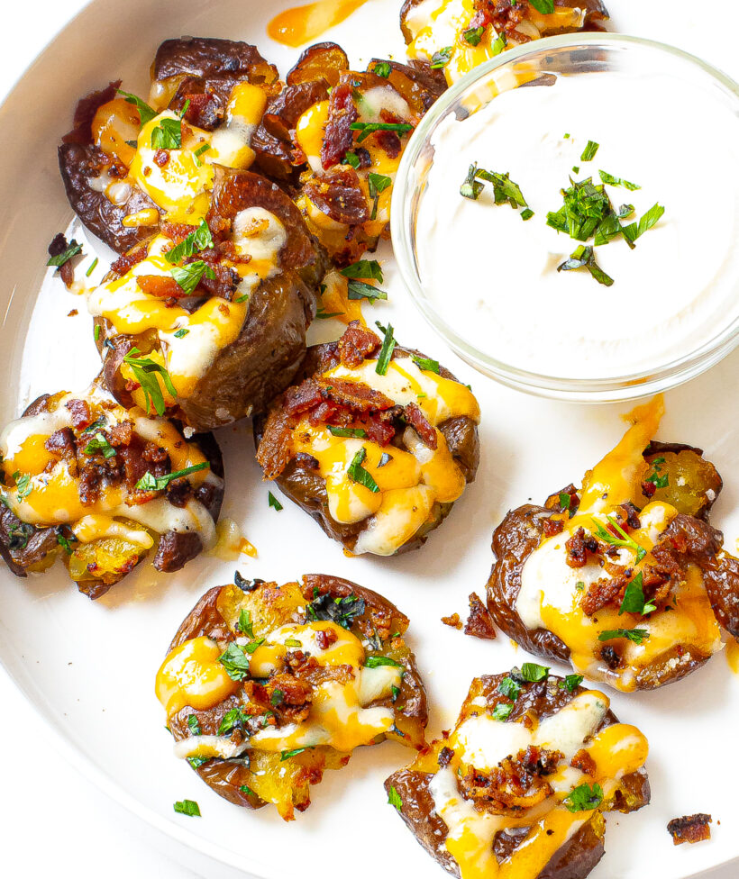 Loaded smashed potatoes on a plate next to a small ramekin of sour cream.