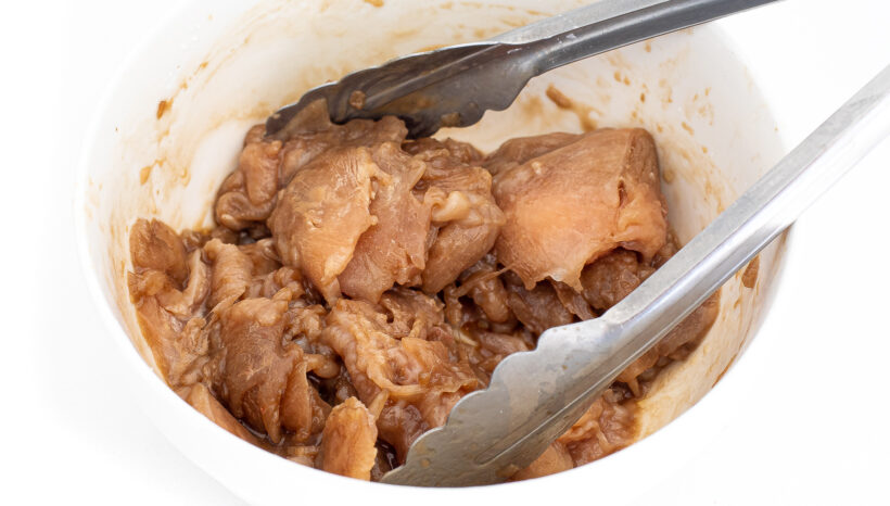 Raw chicken marinating in a bowl with tongs. 