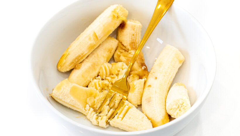 Bananas being mashed in a white bowl with a gold fork.