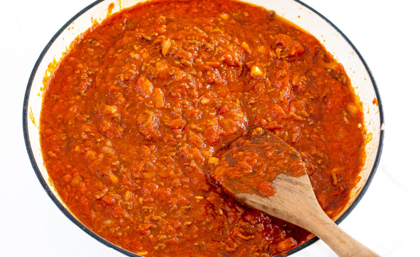 Tomato sauce added to the ground beef mixture in a white skillet with a wooden spoon stirring it.