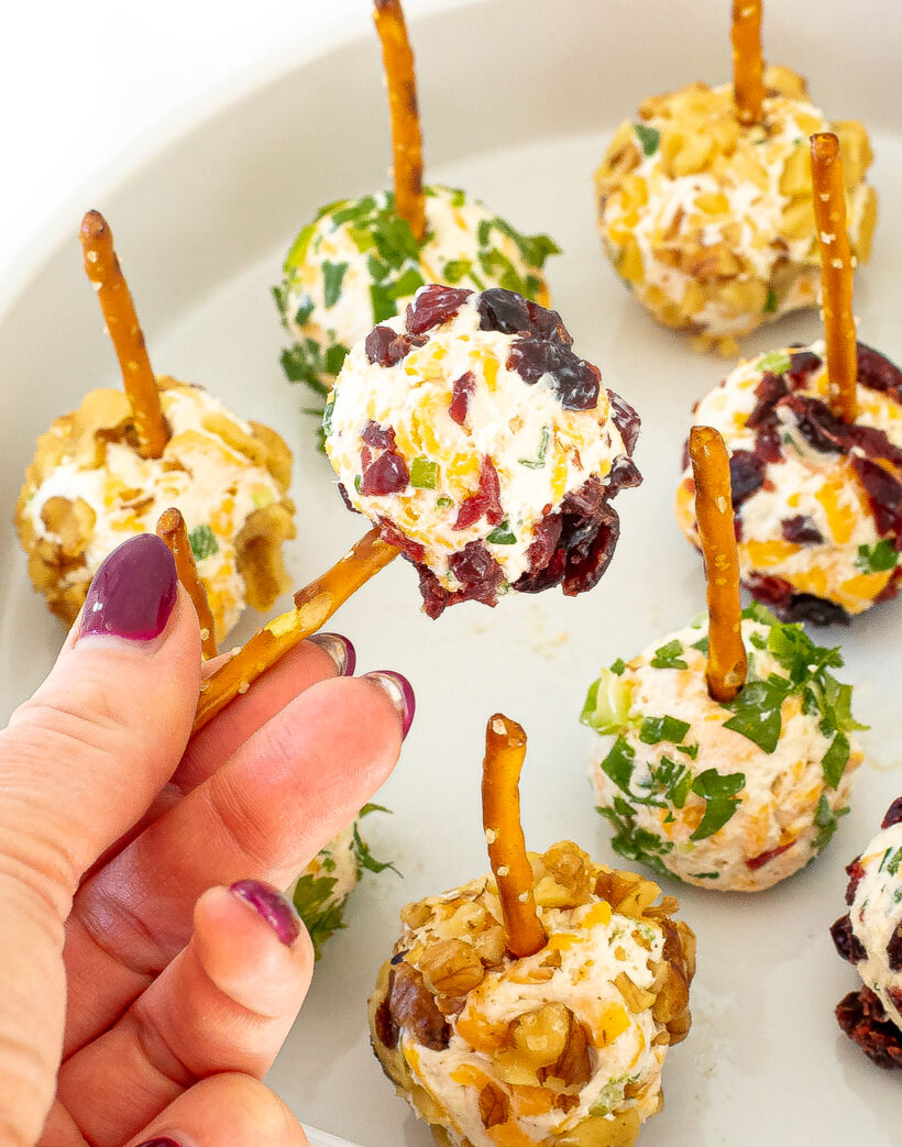 A hand holding up a cranberry cheese ball with a pretzel. There are more cheese balls on a white plate below.